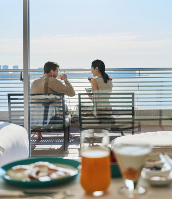写真：客室のテーブルの上には朝食が並び、バルコニー席で朝食を楽しむカップル